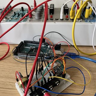 A photo of an Arduino and a breadboard in front of a small modular synth rack connected by a lot of cables
