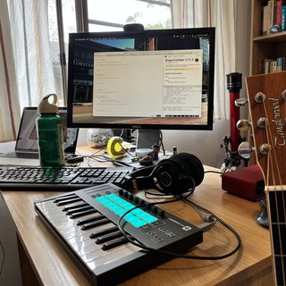 A cluttered desk with a laptop, a monitor, a small MIDI keyboard controller and a pair of headphones. The neck of a Tanglewood guitar is in the foreground.
